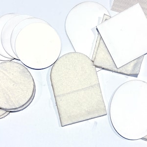 May include: An assortment of white felt and paper shapes, including circles, squares, and rounded rectangles, are scattered on a white surface. The shapes vary in size and texture, with some appearing to be made of a soft, fibrous material.