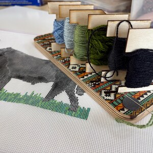 May include: A wooden thread organizer with spools of blue, gray, green, and black thread. The organizer has a decorative patterned design. A cross-stitch project of a gray animal on white fabric is in the foreground.