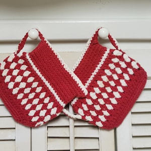 May include: Two red and white crocheted pot holders with a diamond pattern. Each pot holder has a white border and a loop for hanging. The pot holders are hanging on a white surface.