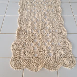 May include: Cream-coloured, open-weave crochet table runner. The runner has a scalloped edge and a repeating circular pattern. The handmade textile is displayed on a white tiled surface.