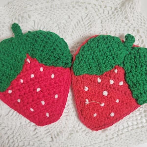 Puede incluir: Dos posavasos hechos a mano en forma de fresa de ganchillo. Los posavasos presentan un cuerpo rojo brillante con detalles de semillas blancas y un tallo verde. Se colocan sobre una superficie blanca texturizada.