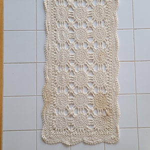May include: Cream-colored, rectangular crochet table runner with a repeating floral pattern. The edges are scalloped, and the runner is displayed on a white tiled surface. The handmade textile adds a touch of vintage charm.