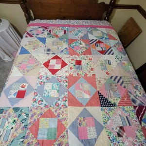 May include: A patchwork quilt featuring a variety of fabric squares in floral, plaid, and solid patterns arranged in a diamond-in-square design. The quilt is bordered by a solid pink fabric strip and displays a diverse mix of colors.