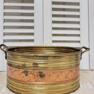 Pode incluir: Um vaso vintage de latão e cobre com duas pequenas alças. O vaso tem uma faixa de cobre com um padrão de círculos repetidos. O fundo apresenta persianas brancas e uma toalha de renda.