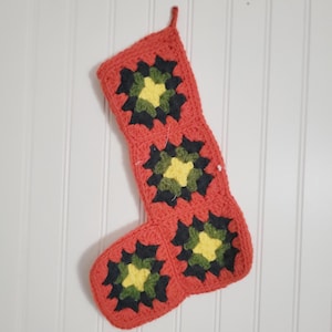 May include: A crochet Christmas stocking constructed from three square granny motifs. Each square features a central yellow flower-like pattern surrounded by dark green and black yarn, all bordered by a vibrant orange yarn frame with a hanging loop.