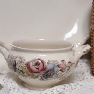 May include: Cream-colored ceramic bowl with two handles, adorned with a floral pattern in pink, purple, and blue. The bowl sits on a white lace doily, with a white plate in the background. A charming piece for serving.