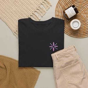 May include: A black short-sleeved t-shirt featuring a small, embroidered starburst design on the left chest. The starburst consists of alternating light purple and dark purple petals arranged in a symmetrical, radiating pattern.