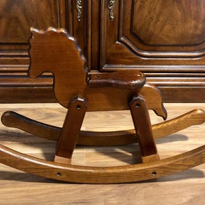 May include: A wooden rocking horse featuring a dark brown finish, a contoured seat, and two curved rockers. The horse has a stylised head with a carved mane and is constructed with visible joints connecting the legs to the body and the rockers.