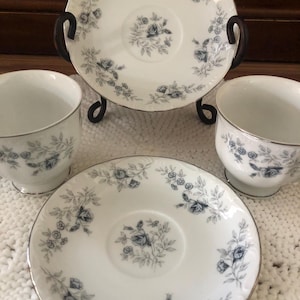 May include: A set of white porcelain teacups and saucers with a blue floral design and gold trim. The teacups and saucers are arranged on a white crocheted surface. The teacups have a rounded shape and sit on a small foot.