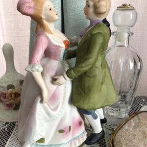 May include: A porcelain figurine depicting a couple in 18th-century attire. The woman wears a pink and white dress, and the man is in a green coat. The figurine is a decorative item, likely for display.