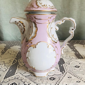 Könnte beinhalten: Eine Vintage-Kaffeekanne aus Porzellan in einem zarten Rosa- und Weißton, verziert mit goldenen Akzenten. Die Kanne hat einen Deckel, einen geschwungenen Griff und aufwendige Details, die ein klassisches, elegantes Design zeigen. Sie steht auf einer Spitzendecke.