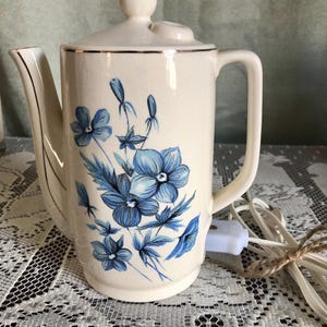 May include: Cream-colored electric coffee pot with a lid and handle. The pot is decorated with blue floral designs and gold trim. A white electrical cord is attached to the pot.