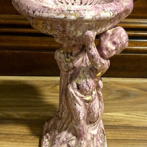 May include: A ceramic pedestal bowl featuring a sculpted figure of a child holding the basin aloft. The piece is finished in a mottled pink and white glaze with gold-toned veining throughout, set upon a square base with a textured, crackled surface.