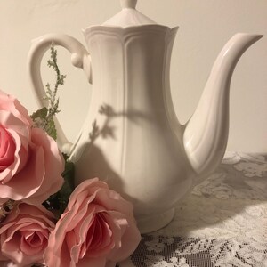 May include: A white porcelain teapot with a fluted body, a matching lid with a finial, a curved handle, and a long spout. The teapot is positioned next to a cluster of artificial light pink roses with green leaves and stems.