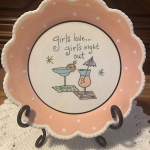 Puede incluir: Un plato decorativo con borde festoneado y diseño de lunares rosas y blancos. El plato presenta el texto "Girls love... girl's night out" e ilustraciones de cócteles. El plato se exhibe sobre un soporte de metal negro.