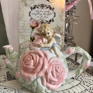 Puede incluir: Una tetera de porcelana blanca con detalles de rosas rosas y una figura de querubín en la tapa. La tetera tiene un asa curva y un pico. El fondo incluye un letrero decorativo con el texto "He has made everything beautiful in its time."
