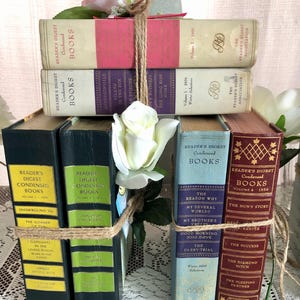 Może przedstawiać: Stos starych książek Reader's Digest Condensed Books związanych sznurkiem i ozdobionych białą różą. Książki mają różne kolory, w tym różowy, fioletowy, niebieski i bordowy, ze złotymi literami.