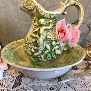 May include: A vintage green ceramic pitcher and basin set. The pitcher features a detailed relief of figures and floral designs. Two pink roses are attached to the pitcher. The basin is white with a green rim. The set is displayed on a lace tablecloth.