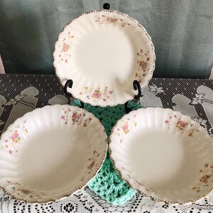 May include: Three white porcelain dishes with a floral pattern and gold trim. The dishes have a scalloped edge. One plate is on a black stand. The floral pattern includes pink, yellow, and purple flowers. The dishes are arranged on a table.