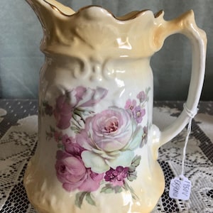 May include: Cream-colored ceramic pitcher with a floral rose design in shades of pink and purple. The pitcher has a scalloped rim and handle, with gold trim accents. A tag is attached to the handle.