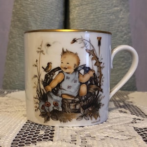 Può includere: Tazza in ceramica bianca con bordo dorato, con un'illustrazione dipinta di un bambino in un cesto con un uccello e fiori. Il bambino indossa un vestito blu. La tazza ha un manico curvo.