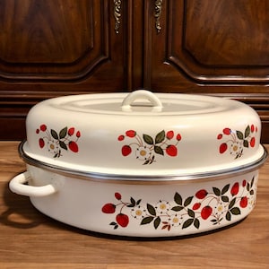 May include: An oval-shaped white enamel roasting pan with a matching lid, featuring a repeating pattern of red strawberries, green leaves, and white blossoms. The pan includes two side handles and a central knob on the lid, with a silver-toned rim.