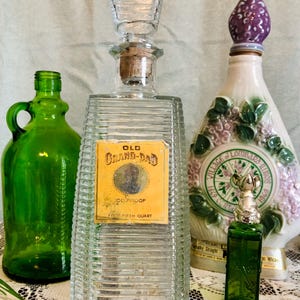 Può includere: Un assortimento di bottiglie di vetro vintage, tra cui una bottiglia rettangolare trasparente con l'etichetta "OLD GRAND-DAD 100 PROOF". Altre bottiglie sono verdi e bianche, con tappi decorativi. Le bottiglie sono disposte su una tovaglia di pizzo.