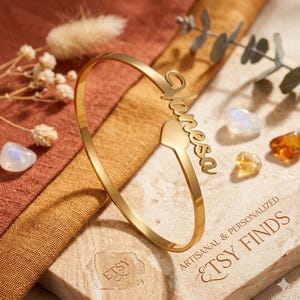 Puede incluir: Una pulsera de puño dorada con el nombre "Vanessa" en escritura cursiva. La pulsera presenta un elemento de diseño en forma de corazón. La imagen está sobre una superficie texturizada con el texto "ARTISANAL & PERSONALIZED ETSY FINDS".