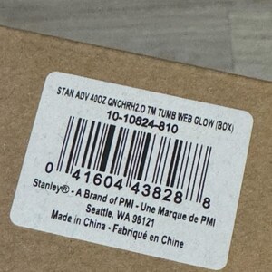 Op de afbeelding: Een wit etiket met zwarte tekst en een barcode. Het etiket luidt "STAN ADV 400Z QNCHRH2.0 TM TUMB WEB GLOW (BOX)" en "Stanley&reg; - A Brand of PMI - Une Marque de PMI". Het etiket bevindt zich op een bruin kartonnen oppervlak.
