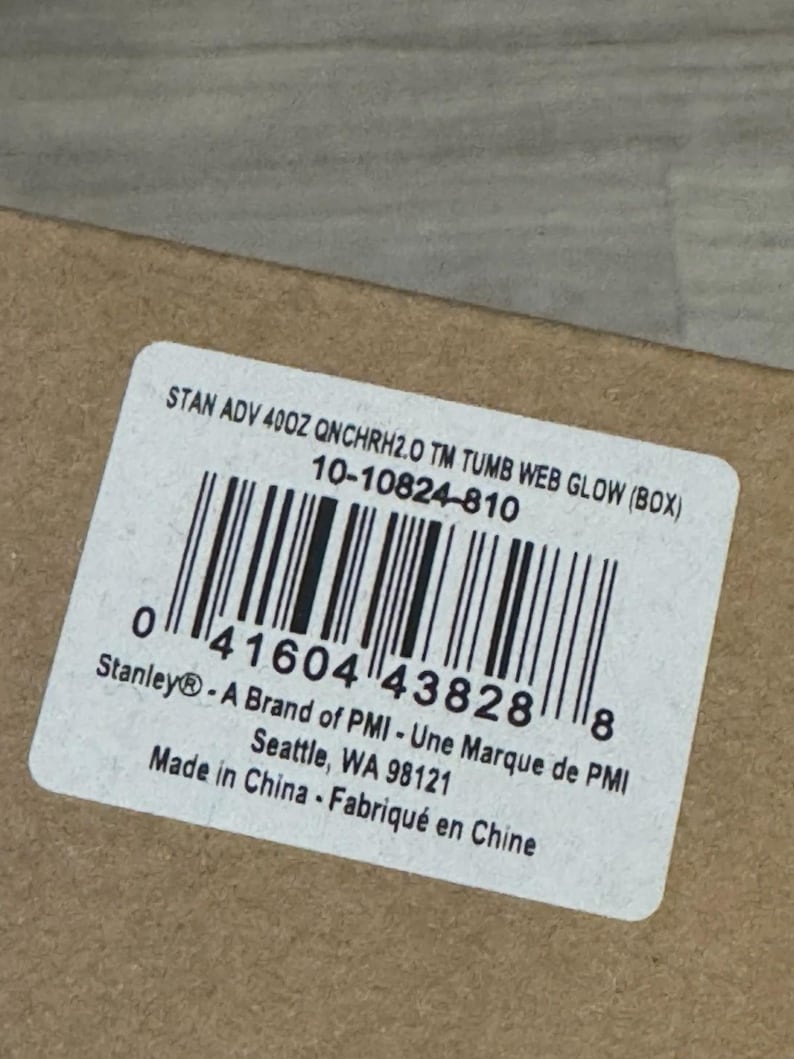 Op de afbeelding: Een wit etiket met zwarte tekst en een barcode. Het etiket luidt "STAN ADV 400Z QNCHRH2.0 TM TUMB WEB GLOW (BOX)" en "Stanley&reg; - A Brand of PMI - Une Marque de PMI". Het etiket bevindt zich op een bruin kartonnen oppervlak.