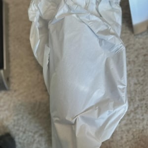 May include: A package wrapped in white tissue paper. The paper is crumpled and folded around an unseen object. The background is a neutral carpet and a glimpse of a metallic object.