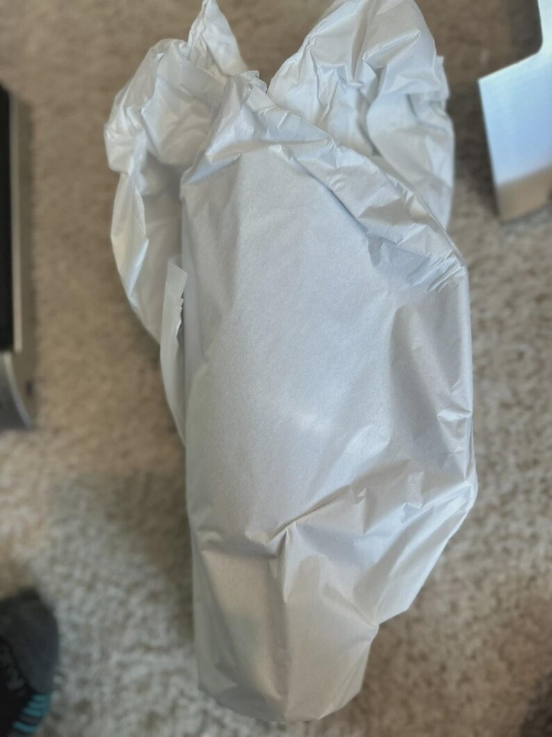 May include: A package wrapped in white tissue paper. The paper is crumpled and folded around an unseen object. The background is a neutral carpet and a glimpse of a metallic object.