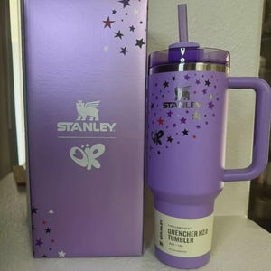 May include: A purple Stanley Quencher H2.0 tumbler with a handle and straw, next to a matching box. The tumbler and box are decorated with star patterns. The tumbler has a 40 oz / 1.18L capacity.