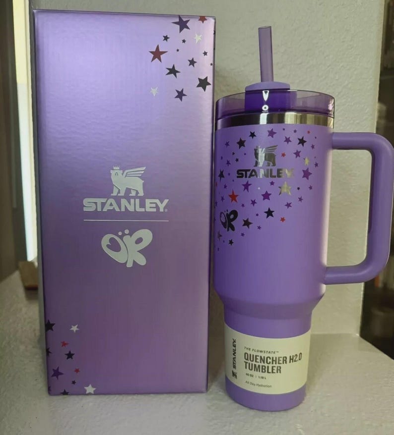 May include: A purple Stanley Quencher H2.0 tumbler with a handle and straw, next to a matching box. The tumbler and box are decorated with star patterns. The tumbler has a 40 oz / 1.18L capacity.