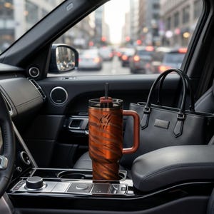 Puede incluir: Un vaso Stanley grande, naranja y marr&oacute;n con un estampado de rayas de tigre y un asa est&aacute; en un coche. El vaso tiene una pajita negra y el logotipo de Stanley. Un bolso negro est&aacute; en el fondo.
