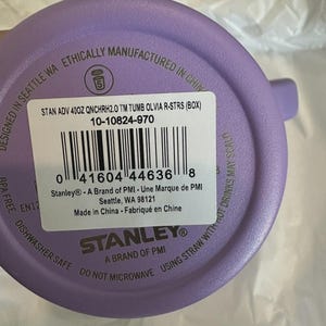 May include: A purple Stanley tumbler with a white label. The label includes the text "STAN ADV 400Z QNCHRH2.0 TM TUMB OLVIA R-STRS (BOX)" and a barcode. The tumbler is dishwasher safe and BPA free.