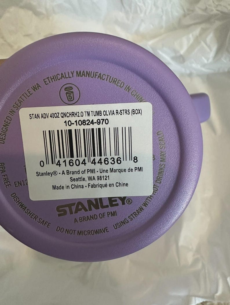May include: A purple Stanley tumbler with a white label. The label includes the text "STAN ADV 400Z QNCHRH2.0 TM TUMB OLVIA R-STRS (BOX)" and a barcode. The tumbler is dishwasher safe and BPA free.