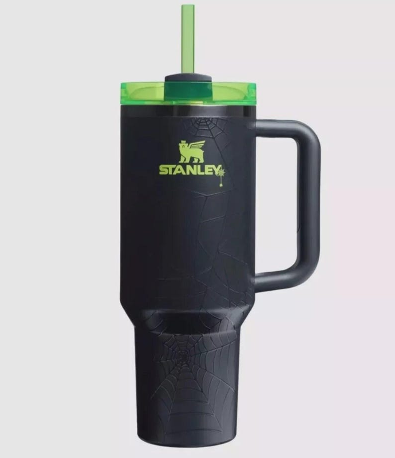 Op de afbeelding: Zwarte Stanley-beker met een groene deksel en rietje. De beker heeft een spinnenwebontwerp en het Stanley-logo in groen. Het heeft een handvat.
