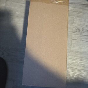 Op de afbeelding: Een rechthoekige kartonnen doos, verzegeld met doorzichtige tape. De doos is lichtbruin van kleur en lijkt leeg te zijn. De doos staat op een houten vloer.