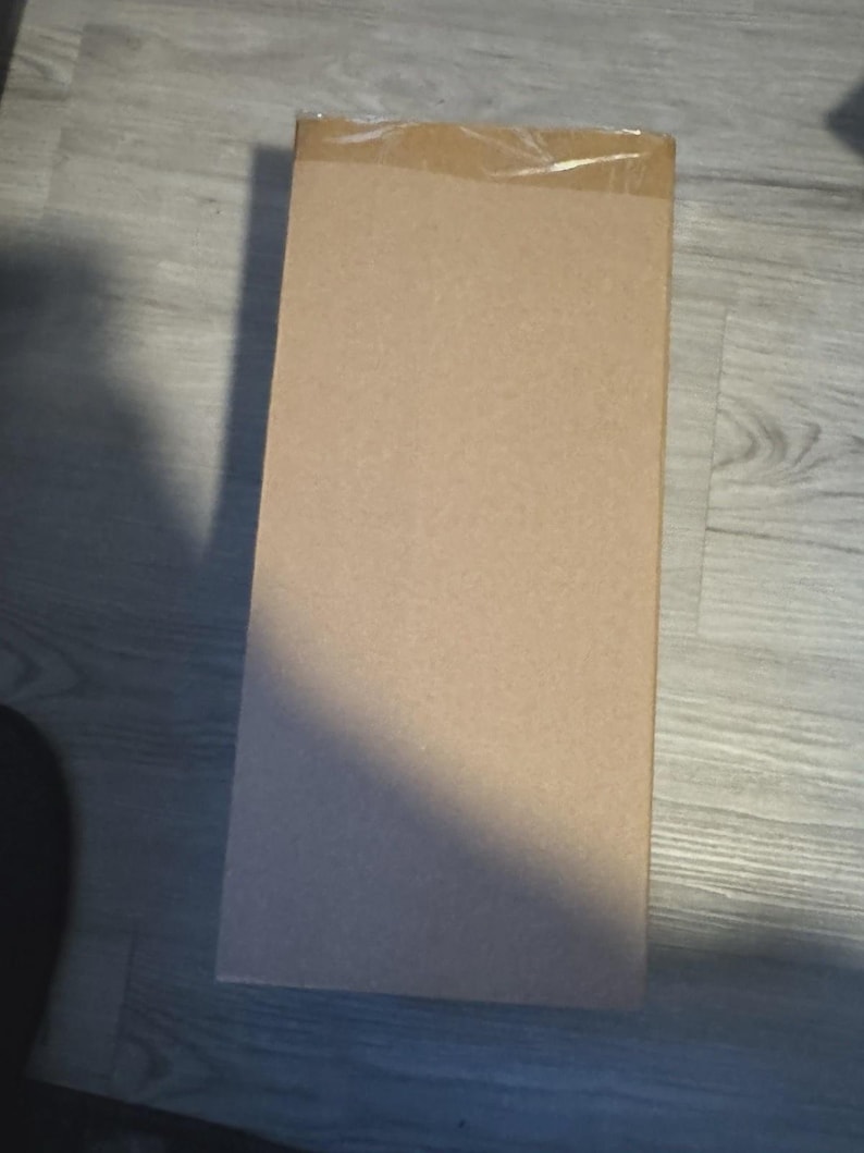 Op de afbeelding: Een rechthoekige kartonnen doos, verzegeld met doorzichtige tape. De doos is lichtbruin van kleur en lijkt leeg te zijn. De doos staat op een houten vloer.