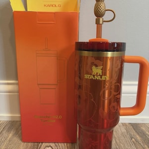 May include: A metallic orange Stanley Quencher H2.0 tumbler featuring a floral pattern, a gold-toned pineapple straw topper, and an orange handle. The tumbler is displayed next to its matching orange and yellow packaging box labeled Stanley and Karol G.