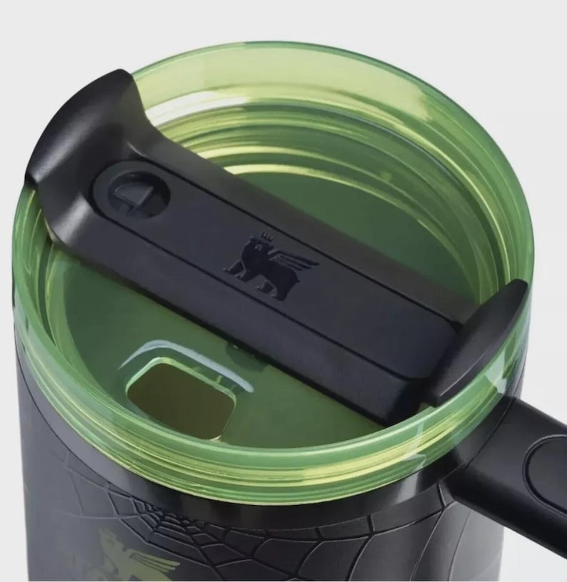 Op de afbeelding: Close-up van een zwart-groene reismok. Het deksel is groen en de schuifregelaar is zwart met een berenlogo. De mok heeft een zwarte buitenkant met een textuurpatroon. De mok is ontworpen voor warme of koude dranken.