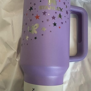 May include: A lavender Stanley Quencher H2O tumbler with a handle and straw. The tumbler is decorated with silver and black stars and a butterfly. The label reads "40 oz / 1.18 L".