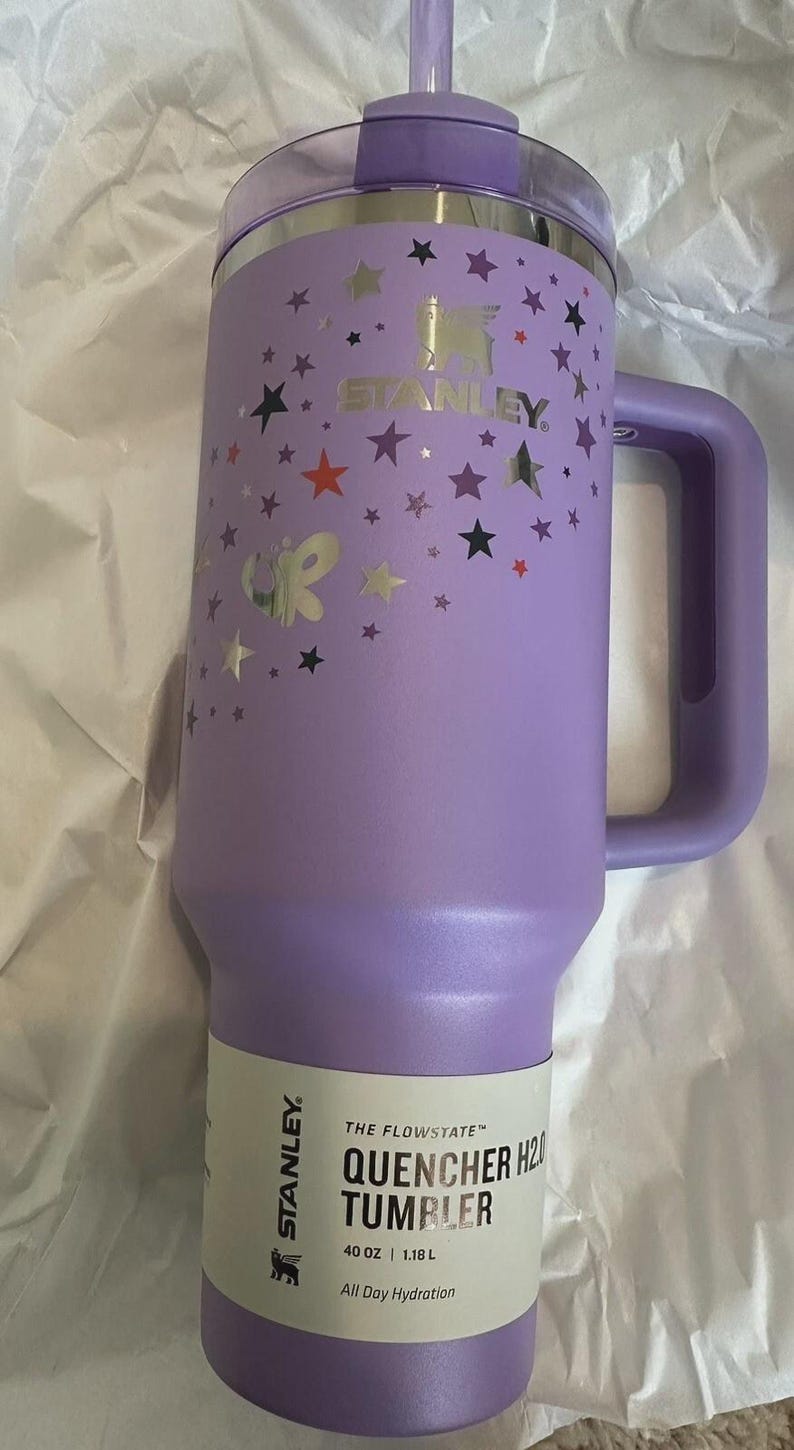 May include: A lavender Stanley Quencher H2O tumbler with a handle and straw. The tumbler is decorated with silver and black stars and a butterfly. The label reads "40 oz / 1.18 L".