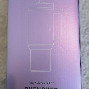 May include: A lavender box featuring a line drawing of a tumbler with a handle and straw. The text reads "THE FLOWSTATE QUENCHER H2.0 TUMBLER" and indicates a 40 oz capacity.