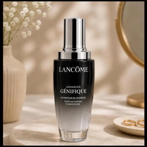 May include: A black and silver Lanc&ocirc;me Advanced G&eacute;nifique Youth Activating Concentrate bottle. The bottle has a gradient design, transitioning from black to clear. The product name and details are printed in white. The background includes a vase with white flowers and a gold-framed mirror.