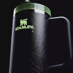 Op de afbeelding: Zwarte Stanley-mok met een groene deksel en rietje. De mok heeft een textuurpatroon en het Stanley-logo in groen. De handgreep is zwart en de achtergrond is donker.