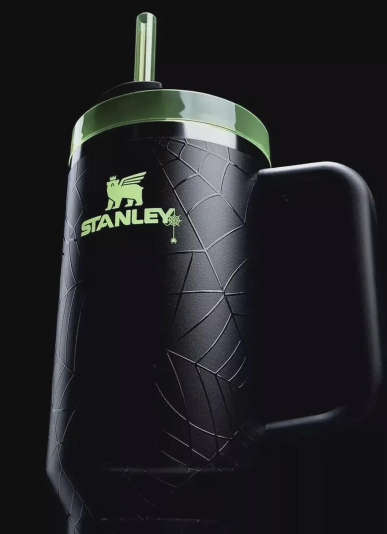Op de afbeelding: Zwarte Stanley-mok met een groene deksel en rietje. De mok heeft een textuurpatroon en het Stanley-logo in groen. De handgreep is zwart en de achtergrond is donker.