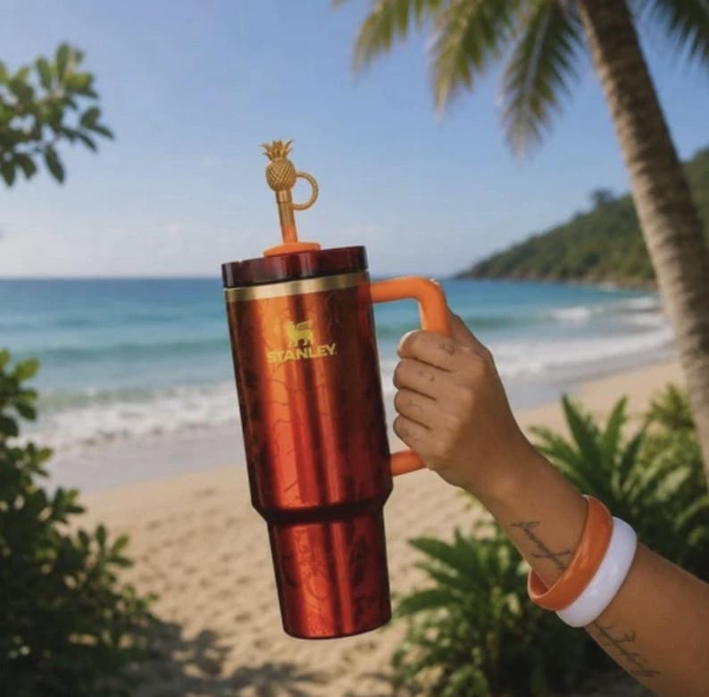 May include: A metallic orange Stanley tumbler with a handle and a gold-colored pineapple-shaped straw topper. The cylindrical cup features the Stanley logo in gold lettering on the front, a dark lid, and a matching orange straw extending from the top.