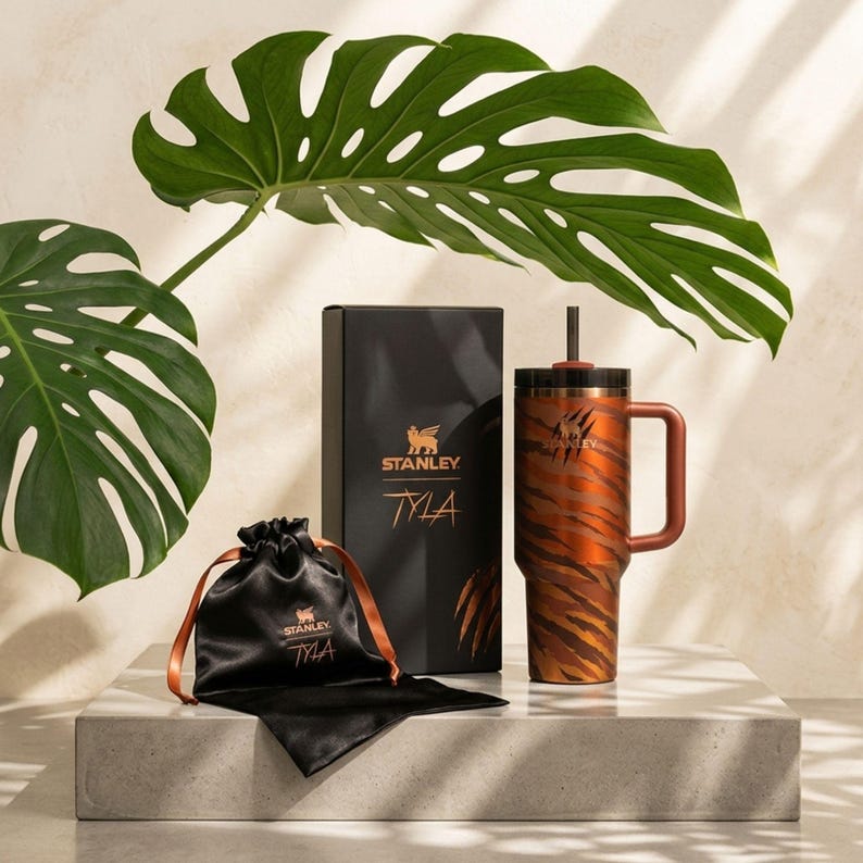 Puede incluir: Un vaso de acero inoxidable con dise&ntilde;o de rayas de tigre, asa marr&oacute;n y tapa negra con pajita. El vaso forma parte de un conjunto que incluye una caja negra y una bolsa negra con cord&oacute;n. La caja y la bolsa tienen el texto "TYLA".